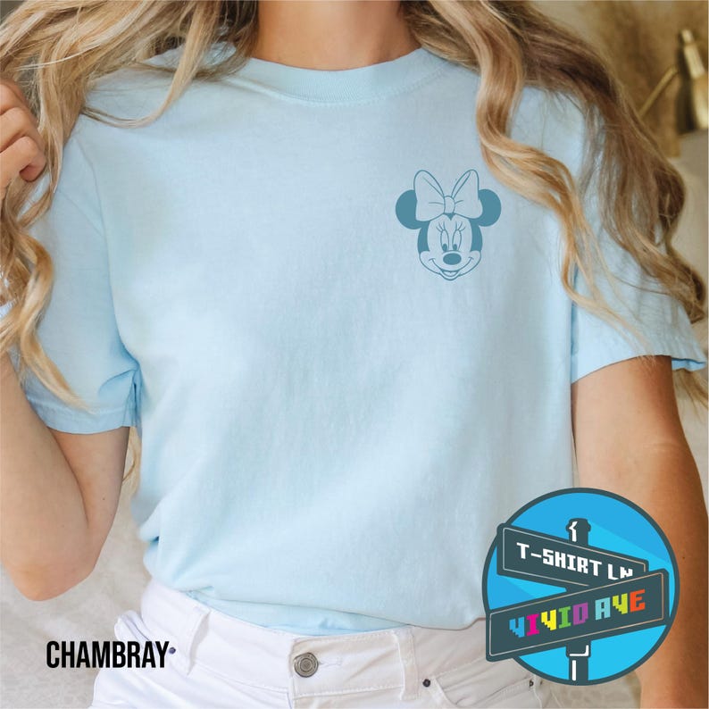 May include: A light blue t-shirt with a small, blue outline of Minnie Mouse's face on the left chest. The t-shirt is on a model wearing white jeans. The text "T-Shirt Ln" and "Vivid Ave" is on a blue sign with rainbow letters.