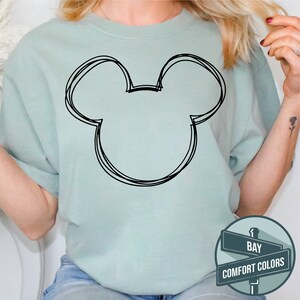 Puede incluir: Una camiseta verde claro con un contorno negro de la cabeza de Mickey Mouse. La camiseta es de color liso y tiene un corte holgado. El letrero en la esquina inferior derecha dice "BAY COMFORT COLORS."