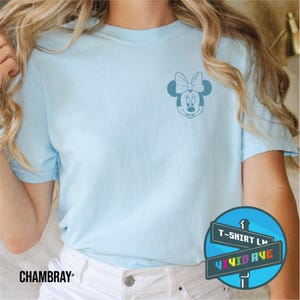May include: A light blue t-shirt with a small, blue outline of Minnie Mouse's face on the left chest. The t-shirt is on a model wearing white jeans. The text "T-Shirt Ln" and "Vivid Ave" is on a blue sign with rainbow letters.
