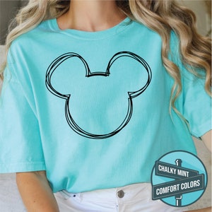 Puede incluir: Una camiseta azul claro con un contorno negro de la cabeza de Mickey Mouse. La camiseta es de color liso y tiene cuello redondo. La imagen tambi&eacute;n incluye un letrero que dice "Chalky Mint" y "Comfort Colors."