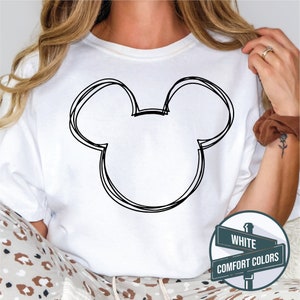 Puede incluir: Camiseta blanca con un contorno negro de la cabeza de Mickey Mouse. El dise&ntilde;o es de estilo sencillo, dibujado a mano. La camiseta forma parte de la colecci&oacute;n "Comfort Colors".