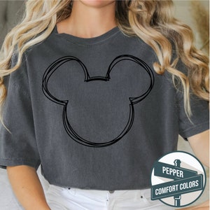 Puede incluir: Camiseta gris oscuro con un contorno negro de la cabeza de Mickey Mouse. La camiseta tiene cuello redondo y mangas cortas. El texto en el letrero dice "Pepper Comfort Colors".