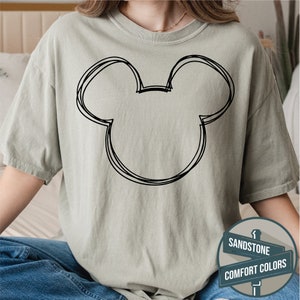 Puede incluir: Camiseta color arena con un contorno negro de la cabeza de Mickey Mouse. El dise&ntilde;o es de estilo simple y esbozado. La camiseta forma parte de la colecci&oacute;n Comfort Colors.