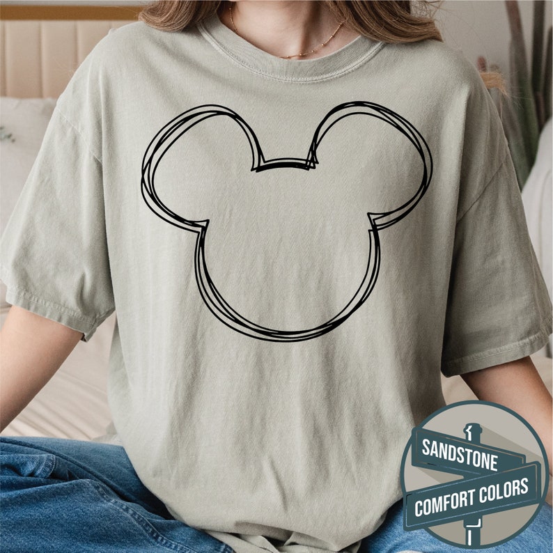 Puede incluir: Camiseta color arena con un contorno negro de la cabeza de Mickey Mouse. El dise&ntilde;o es de estilo simple y esbozado. La camiseta forma parte de la colecci&oacute;n Comfort Colors.
