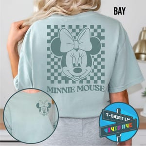 May include: A light blue t-shirt with a checkered pattern and a gray Minnie Mouse graphic on the back. The text "Minnie Mouse" is printed below the graphic. The t-shirt has a crew neck and short sleeves.