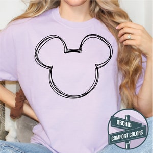 Puede incluir: Una camiseta de color lila claro con un contorno negro de la cabeza de Mickey Mouse. El dise&ntilde;o es de estilo sencillo, dibujado a mano. La camiseta es de color liso, con las palabras "ORCHID" y "COMFORT COLORS" en un letrero.
