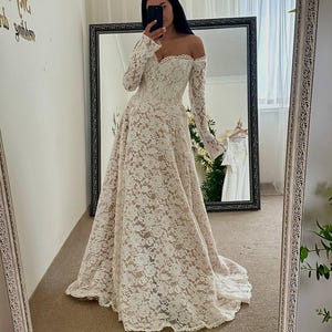 May include: Off-the-shoulder ivory lace wedding dress with long sleeves and a flowing skirt. The dress features a sweetheart neckline and a delicate floral lace pattern. The bride is taking a mirror selfie.