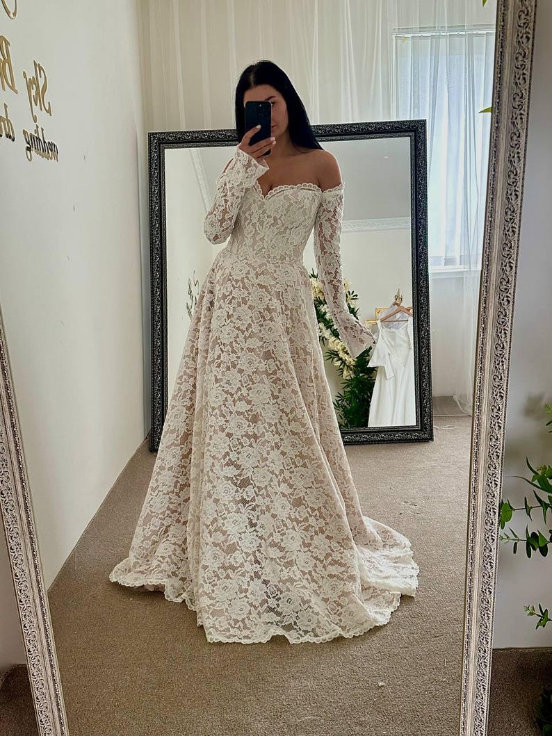 May include: Off-the-shoulder ivory lace wedding dress with long sleeves and a flowing skirt. The dress features a sweetheart neckline and a delicate floral lace pattern. The bride is taking a mirror selfie.
