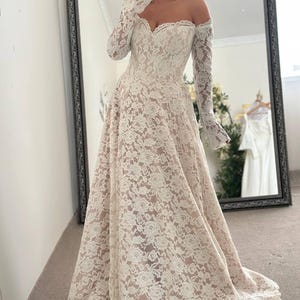 May include: Off-the-shoulder ivory lace wedding dress with long sleeves. The dress features a sweetheart neckline and a full skirt. The intricate lace detailing covers the entire gown, creating a romantic and elegant look. The dress has a slight train.