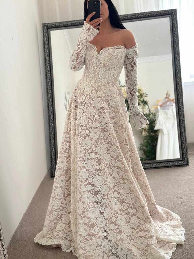 May include: Off-the-shoulder ivory lace wedding dress with long sleeves. The dress features a sweetheart neckline and a full skirt. The intricate lace detailing covers the entire gown, creating a romantic and elegant look. The dress has a slight train.