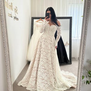 May include: A white lace wedding dress with long sleeves and a sweetheart neckline. The dress has a full skirt and a train. The image is taken in front of a mirror, with other dresses visible in the background.