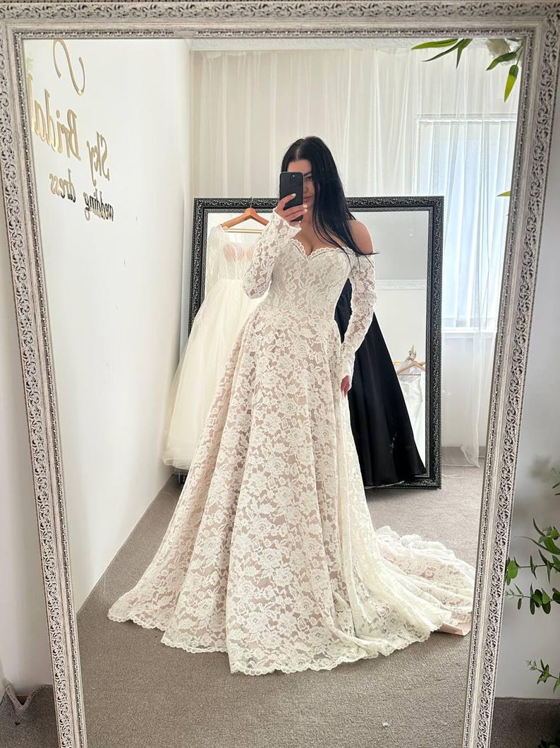May include: A white lace wedding dress with long sleeves and a sweetheart neckline. The dress has a full skirt and a train. The image is taken in front of a mirror, with other dresses visible in the background.