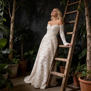May include: A white lace off-the-shoulder wedding dress with long sleeves. The dress features a floral pattern and a flowing skirt. A wooden ladder is in the background, surrounded by potted plants and trees.