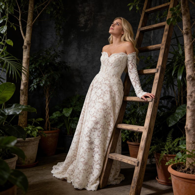 May include: A white lace off-the-shoulder wedding dress with long sleeves. The dress features a floral pattern and a flowing skirt. A wooden ladder is in the background, surrounded by potted plants and trees.