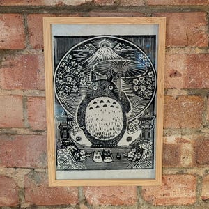 May include: Framed black and white print featuring a large, round, furry creature with a white belly, set against a backdrop of a mountain, flowers, and a large umbrella. The print is in a light brown wooden frame.