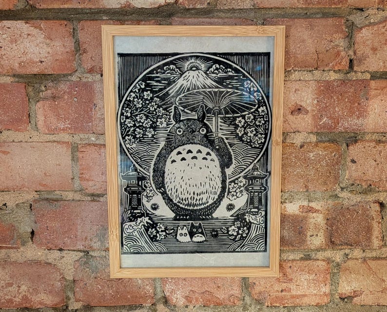 May include: Framed black and white print featuring a large, round, furry creature with a white belly, set against a backdrop of a mountain, flowers, and a large umbrella. The print is in a light brown wooden frame.