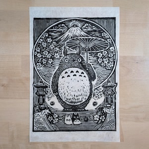 May include: Black and white woodcut print featuring a large, round creature holding an umbrella, with a mountain and cherry blossoms in the background. Two smaller creatures are at the bottom. Japanese-inspired art.