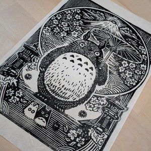 May include: Black and white linocut print featuring a large, round, furry creature holding an umbrella. The print includes Japanese-style imagery of a mountain, flowers, and pagodas. The artwork is on a light-colored paper.