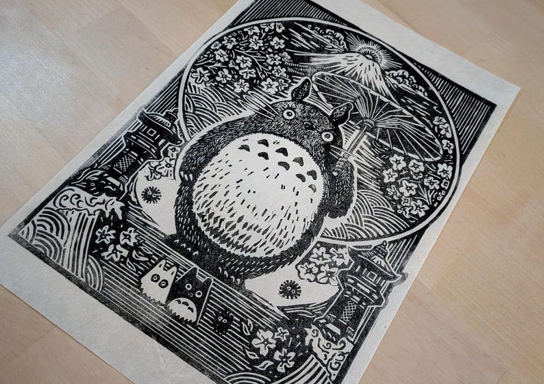 May include: Black and white linocut print featuring a large, round, furry creature holding an umbrella. The print includes Japanese-style imagery of a mountain, flowers, and pagodas. The artwork is on a light-colored paper.