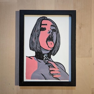 May include: Framed pop art print depicting a woman with grey hair, pink skin, and a black collar. The woman has her mouth open with her tongue out, and she is pointing at the collar. The artwork is in a black frame.