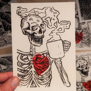 Puede incluir: Una ilustración en blanco y negro de un esqueleto sosteniendo una taza de café humeante. El esqueleto tiene un corazón rojo en su pecho.