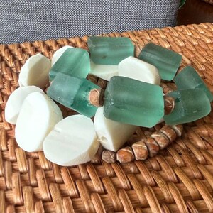 May include: A handmade bracelet featuring sea glass beads in shades of green and white, interspersed with small, brown, cylindrical beads. The bracelet is displayed on a woven wicker surface.