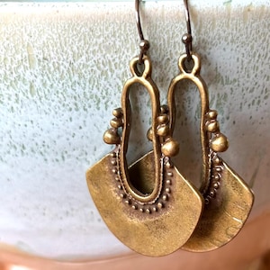 Puede incluir: Un par de pendientes de color latón antiguo. Cada pendiente presenta una base en forma de abanico con un borde punteado y una sección superior curva y abierta adornada con pequeños detalles redondos. Los pendientes cuelgan de un gancho simple.