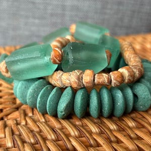 Könnte beinhalten: Drei gestapelte Armbänder mit Seeglas- und Holzperlen. Das obere Armband hat große, rechteckige, seegrüne Glasperlen. Das mittlere Armband hat Holzperlen. Das untere Armband hat abgerundete, teal-farbene Perlen.