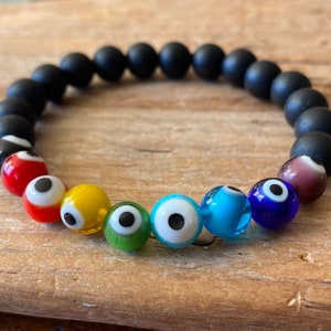 May include: A black beaded bracelet with rainbow colored evil eye charms. The bracelet is made of matte black beads and features seven evil eye charms in red, yellow, green, white, blue, turquoise, and purple.