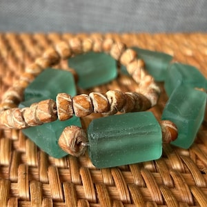 Op de afbeelding: Een handgemaakte armband met grote, rechthoekige zeeglas kralen in een doorschijnende teal kleur. Kleine, lichtbruine houten kralen zijn afgewisseld, geregen op een natuurlijk gekleurd koord. De armband wordt gepresenteerd op een geweven oppervlak.