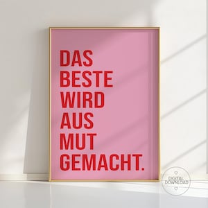 May include: A framed pink print with red text that reads "DAS BESTE WIRD AUS MUT GEMACHT." A small digital download logo is in the bottom right corner. The frame is gold.