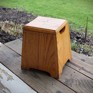 May include: A wooden step stool with a rectangular top and a trapezoidal body. The stool is made of light brown wood with visible grain and has a handle cut out on each side. It is sitting on a wooden deck.