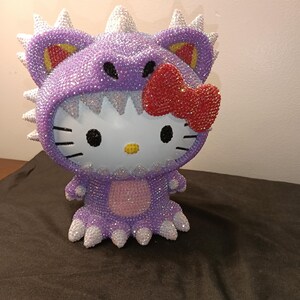 May include: A bedazzled figurine of a cartoon cat in a purple dinosaur costume with white spikes. The cat has a red bow, black eyes, and whiskers. The figurine is covered in sparkling crystals.