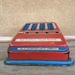 May include: A vintage red and blue adding machine with the text "ADDING MACHINE" on the front. The machine has several blue number sliders and is made by Wolverine Supply & Mfg. Co. in Pittsburgh, PA.