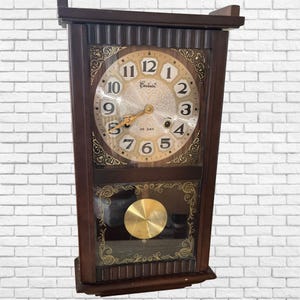 Könnte beinhalten: Eine Vintage-Wanduhr aus Holz mit rechteckiger Form. Das Zifferblatt ist beige mit schwarzen Ziffern und goldfarbenen Zeigern. Die Uhr hat ein Pendel und dekorative goldfarbene Akzente. Die Uhr zeigt "35 DAY" unterhalb der Mitte an.
