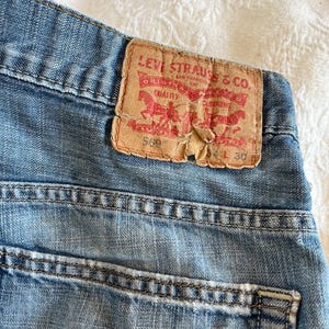 Puede incluir: Primer plano de unos vaqueros de mezclilla azul claro con una etiqueta descolorida de Levi Strauss & Co. La etiqueta es marrón con letras rojas y un gráfico. Los vaqueros tienen una cintura de 76 cm.