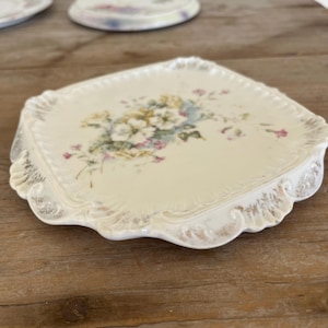 Könnte beinhalten: Ein quadratischer Untersetzer aus weißer Keramik mit einem Blumenmuster. Der Untersetzer hat einen gewellten Rand und eine weiße, abgenutzte Oberfläche.