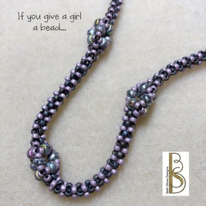 Può includere: Primo piano di una collana di perline di colore viola e nero. La collana presenta un motivo ripetitivo di piccole perline e perline più grandi e raggruppate. Il testo "If you give a girl a bead..." è visibile nell'angolo in alto a sinistra dell'immagine.