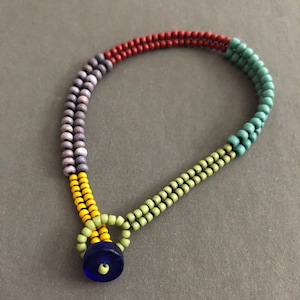 May include: A multi-coloured beaded bracelet with a blue glass bead in the centre. The bracelet is made with red, purple, yellow, green, and blue beads.