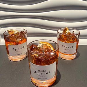 Könnte beinhalten: Drei Aperol-Kerzen in transparenten Gläsern. Jede Kerze hat eine orangefarbene Wachsbasis, gefüllt mit bernsteinfarbenem Wachs und eisartigen Verzierungen sowie einer getrockneten Orangenscheibe. Die Etiketten lauten "Aperol Candle" und "Nicoles Aperol Candle".