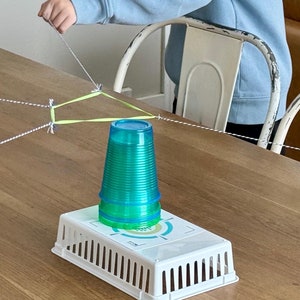 May include: A green plastic cup sits on a white plastic base with a circular design. The cup is held in place by four strings tied to a green and white triangle.