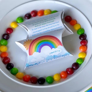 May include: A white plate with a circle of colorful candy around the edge. In the center of the plate are two small pillow boxes with a rainbow, sun, and clouds design. The boxes have the text "3 Nephi 22:7-10" printed on them.