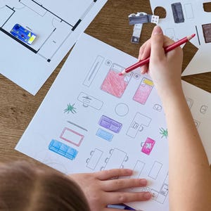 May include: A child colors in a black and white drawing of a house floor plan with a red colored pencil. The drawing includes furniture, a bed, a couch, a table, and a plant.