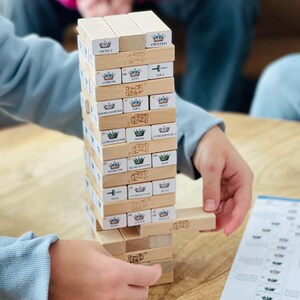 May include: A person playing a game of Jenga, a classic wooden block stacking game. The tower is tall and precariously balanced, with one block being carefully removed. The game is played on a wooden table.