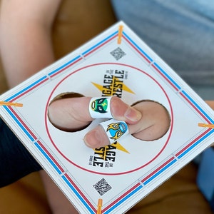 May include: A white cardboard game with blue and red trim. The game is called "Engage in the Finger Wrestle" and has two holes for fingers with wrestling masks on them.
