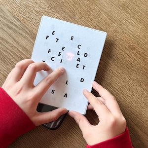 May include: A white card with black letters spelling out words is held over a smartphone. The card is being touched by fingers, and the person is wearing a red long-sleeved shirt. The background is a wooden surface.