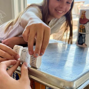 May include: Three small, white, cylindrical objects with black text are being manipulated by hands. The text on the objects reads "WITHOUT HANDS SHALL ROLL FORTH, FORTH, PLEASE". A decorative, painted ceramic object is in the background.
