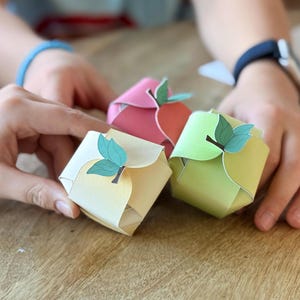 May include: Three colorful, origami-style gift boxes shaped like apples. The boxes are in shades of yellow, pink, and green, each with a leaf and stem detail. The boxes are sitting on a wooden surface.