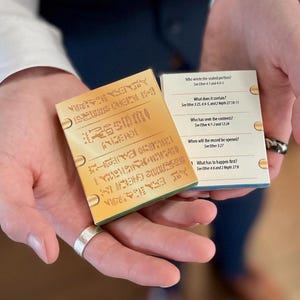 Puede incluir: Dos pequeños libros de color dorado con preguntas sobre el Libro de Éter. Las preguntas están impresas en la parte delantera de los libros. Los libros están en manos de una persona.