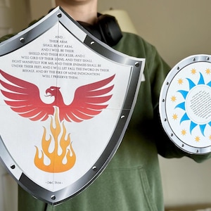 May include: Two cardboard shields with a silver border. The larger shield features a red phoenix with orange flames and a white background. The smaller shield has a blue sun with yellow stars on a white background. Both shields have text on them. The text on the larger shield reads: "AND THEIR ARM SHALL BE MY ARM. AND I WILL BE THEIR SHIELD AND THEIR BUCKLER, AND I WILL GIRD UP THEIR LOINS, AND THEY SHALL FIGHT MANFULLY FOR ME, AND THEIR ENEMIES SHALL BE UNDER THEIR FEET: AND I WILL LET FALL THE SWORD IN THEIR BEHALF, AND BY THE FIRE OF MINE INDIGNATION WILL I PRESERVE THEM. -D&C 35:14-"